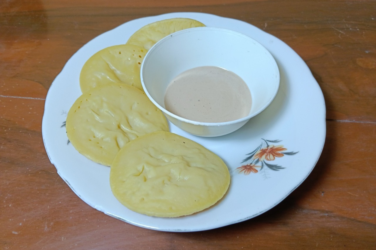 Empat kue apen pipih berwarna kuning pucat di piring bermotif bunga, dengan semangkuk kecil kuah santan di tengahnya