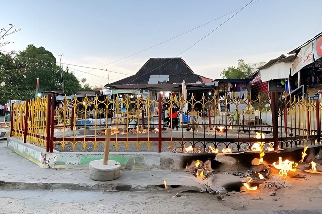 Api menyala di beberapa titik tanah di dalam area berpagar, dengan deretan warung di belakangnya