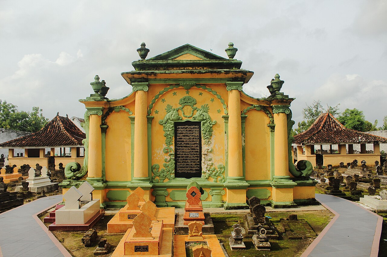 Bangunan makam bercat kuning dengan prasasti hitam di tengah dindingnya