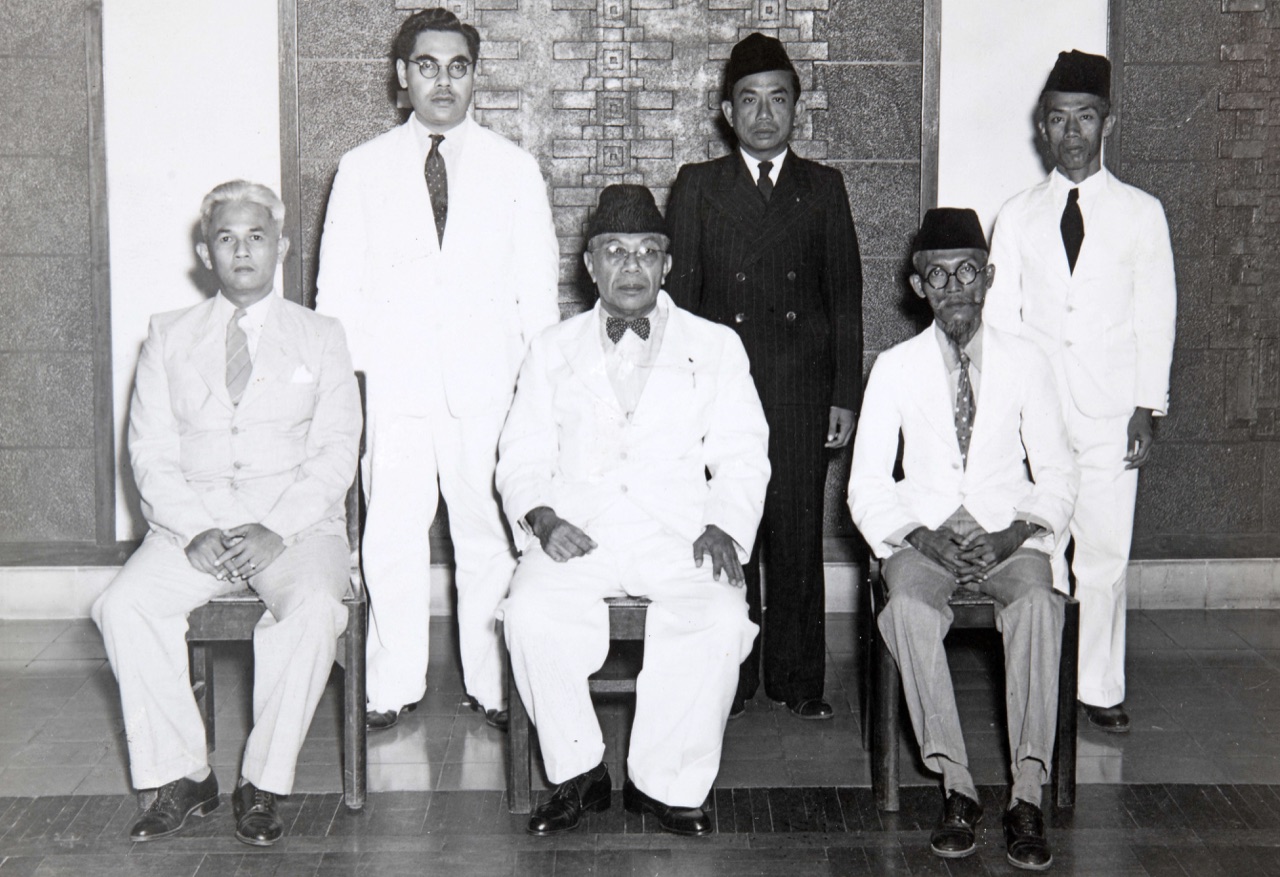 Foto hitam putih enam laki-laki berjas, tiga duduk dan tiga berdiri, dengan Cakraningrat XII berpeci dan berdasi kupu-kupu duduk di tengah