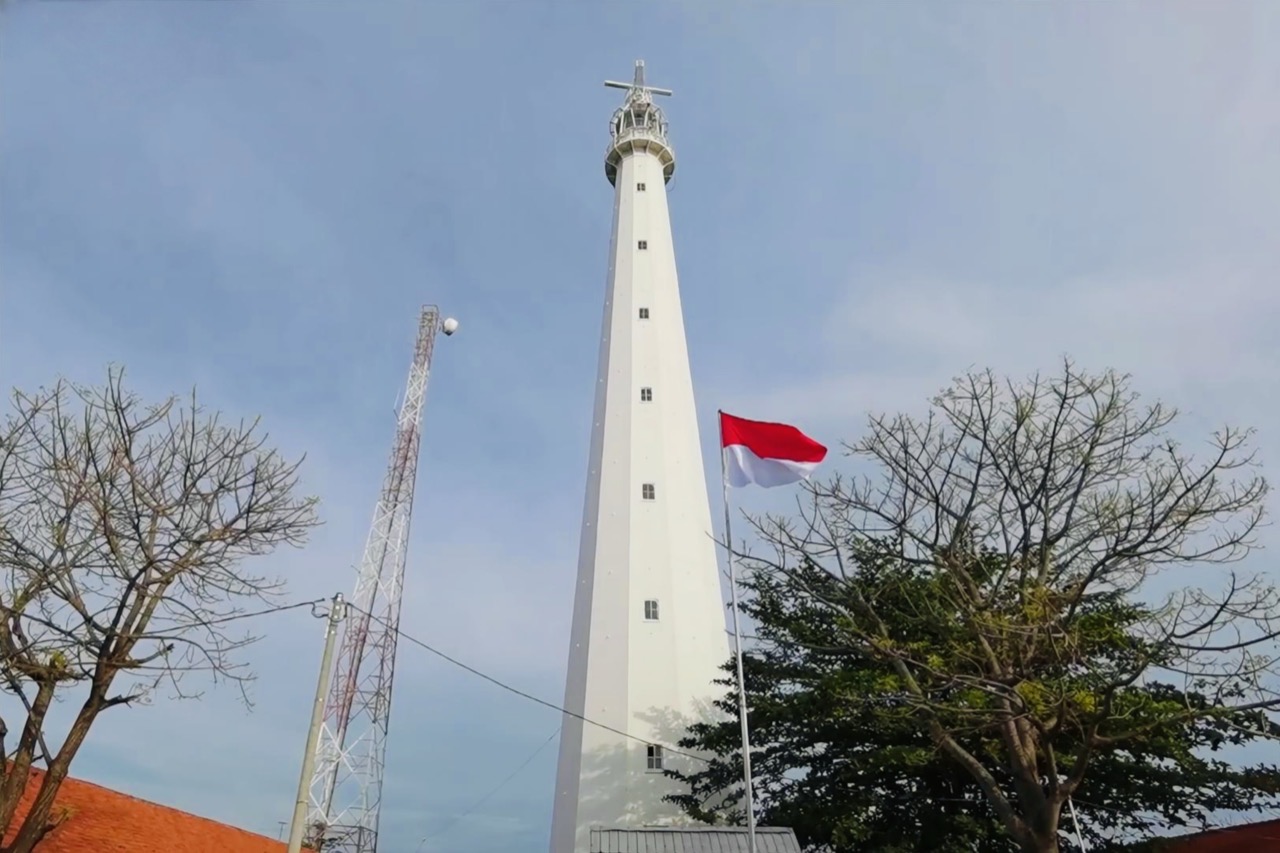 Menara mercusuar besi bercat putih menjulang dilihat dari bawah, bendera Merah Putih berkibar di sebelahnya