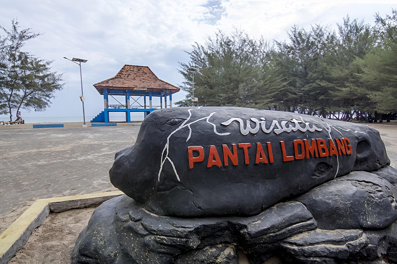 Batu bertuliskan Wisata Pantai Lombang di depan barisan cemara