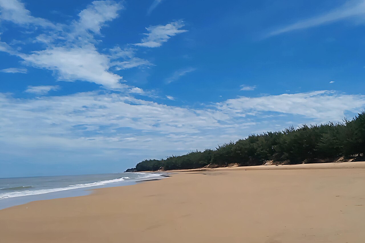 Pantai Slopeng dengan pasir luas, barisan pohon di kejauhan, dan langit berawan
