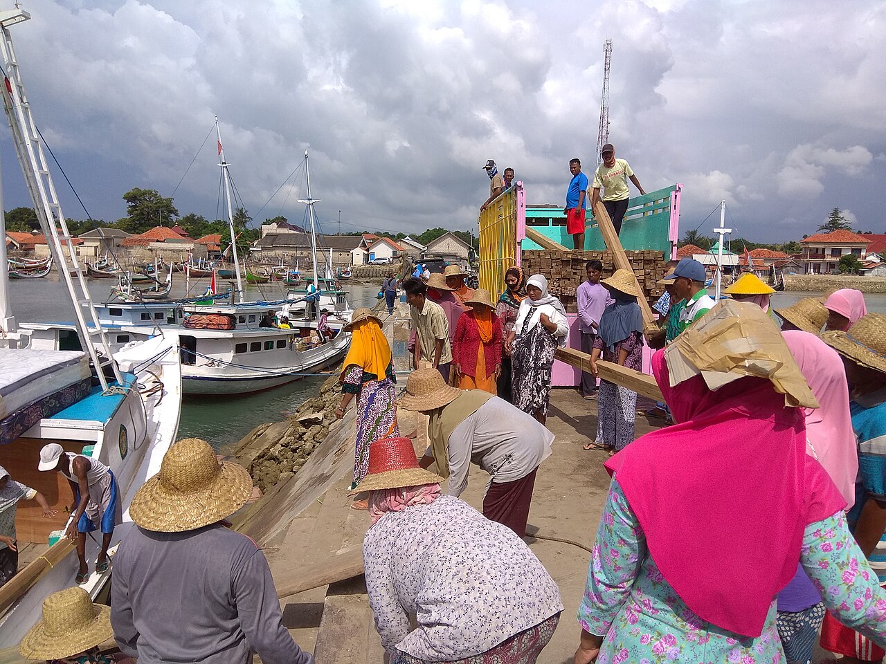 Sekelompok perempuan bertopi lebar memikul barang di pantai berpasir, perahu kayu berjajar di belakangnya