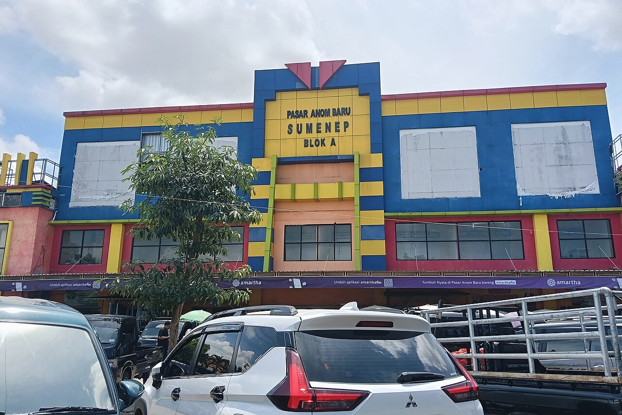 Gedung Pasar Anom Baru Sumenep bercat biru, kuning, dan merah dengan deretan mobil parkir di depannya
