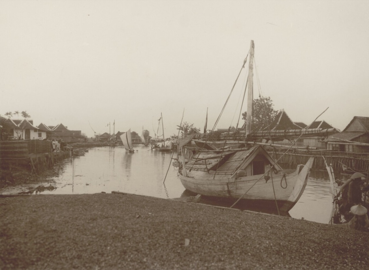 Foto sepia lama memperlihatkan perahu Madura berumah geladak anyaman bersandar di alur sungai berumah panggung