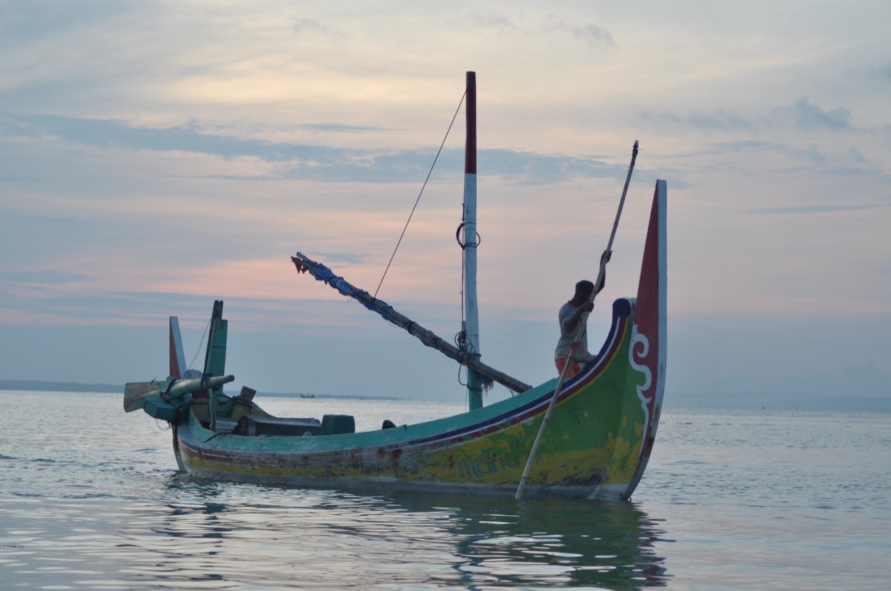 Perahu nelayan berhaluan tinggi bercat hijau, kuning, dan merah dengan sulur putih, terapung di air tenang saat senja