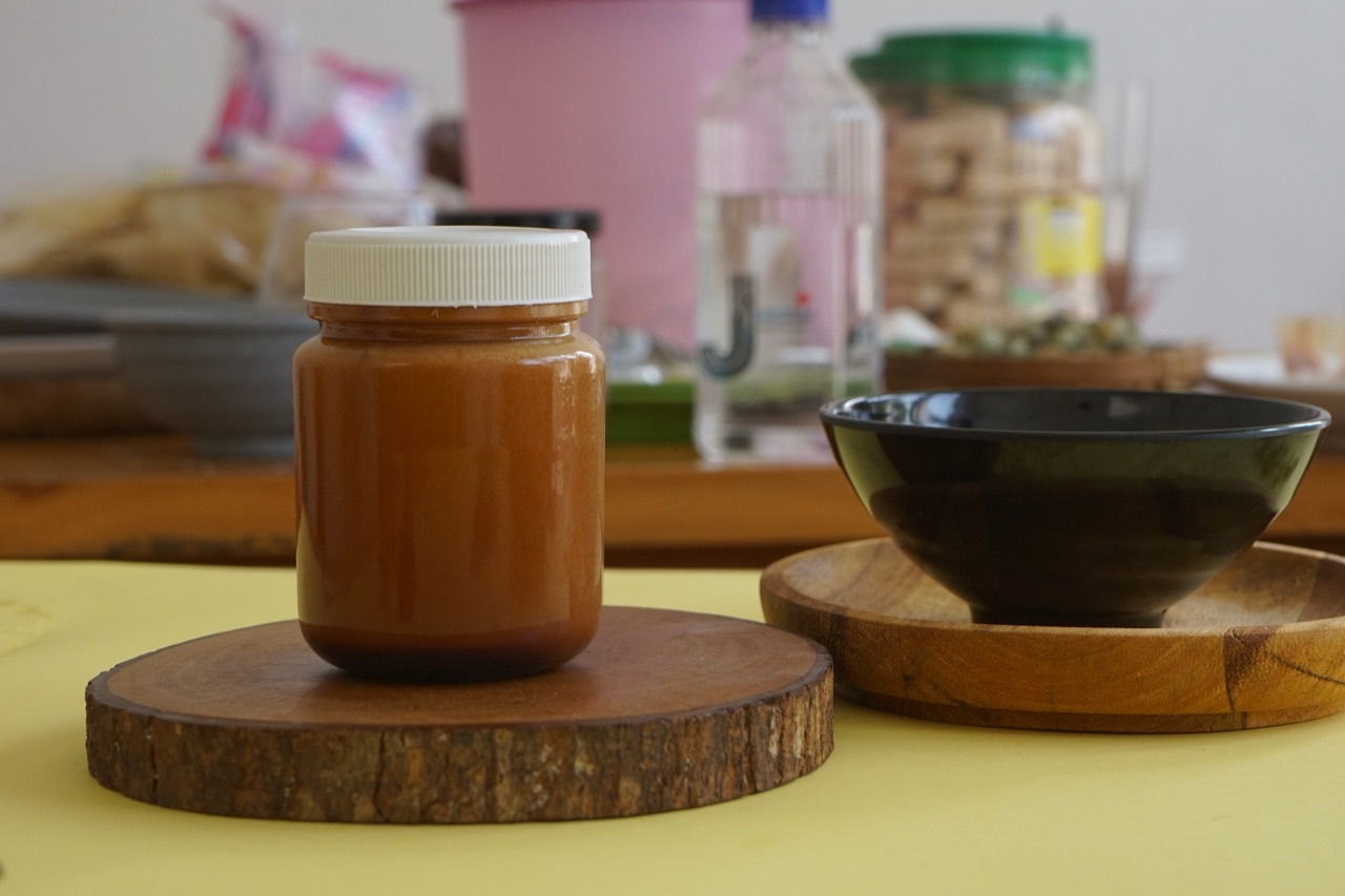 Satu toples kaca berisi petis cokelat kemerahan di atas potongan kayu, dengan mangkuk kecil di sebelahnya