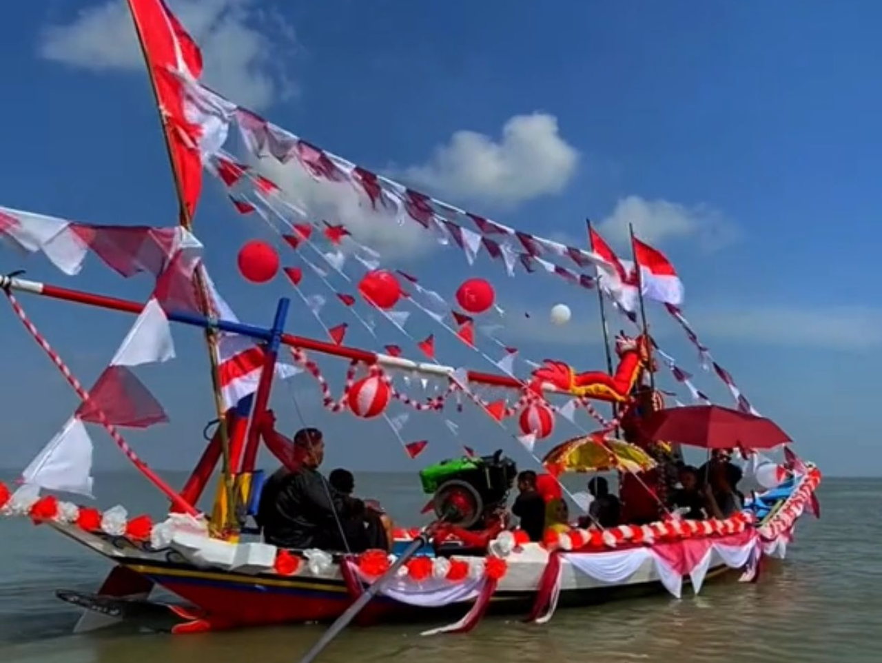 Perahu nelayan berhias bendera merah putih, umbul-umbul, dan balon berlayar di laut dangkal saat Rokat Tase'