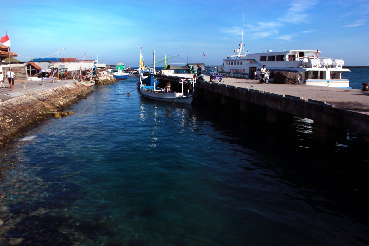Dermaga beton dengan kapal penumpang putih bersandar, perahu kayu di alur air jernih kehijauan di tengahnya