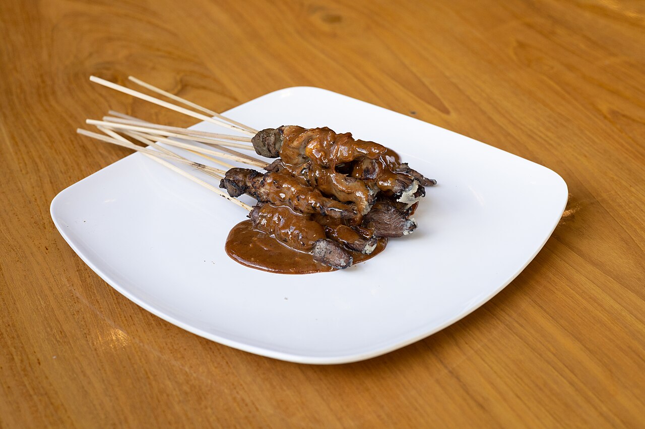 Beberapa tusuk sate berlumur bumbu kacang di piring putih di atas meja kayu