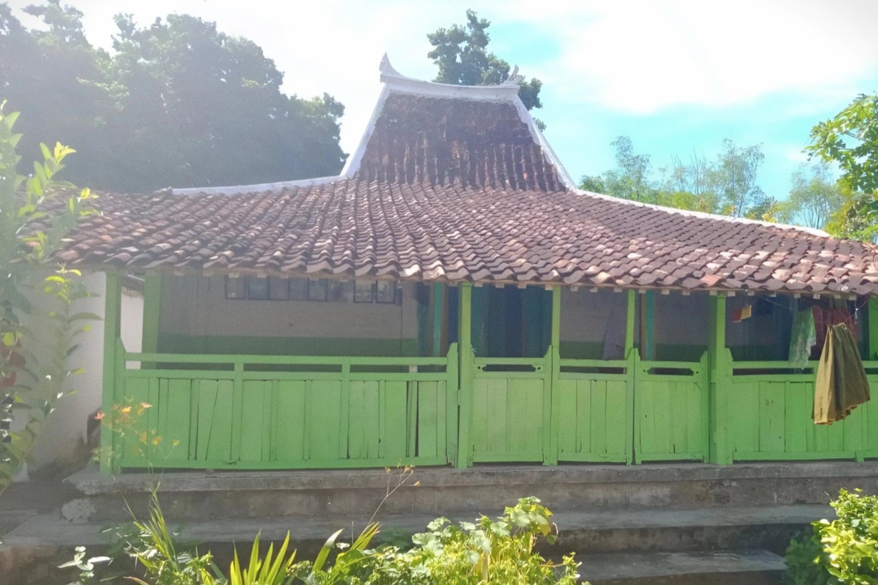 Rumah kayu bercat hijau beratap genteng dengan bubungan melengkung dan serambi berpagar kayu di depannya