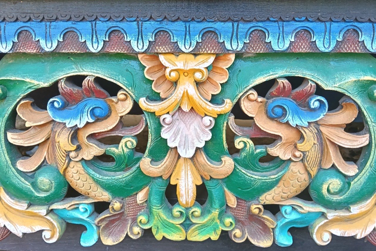 Panel ukiran kayu berwarna dengan dua makhluk bersisik saling berhadapan mengapit motif bunga di tengahnya